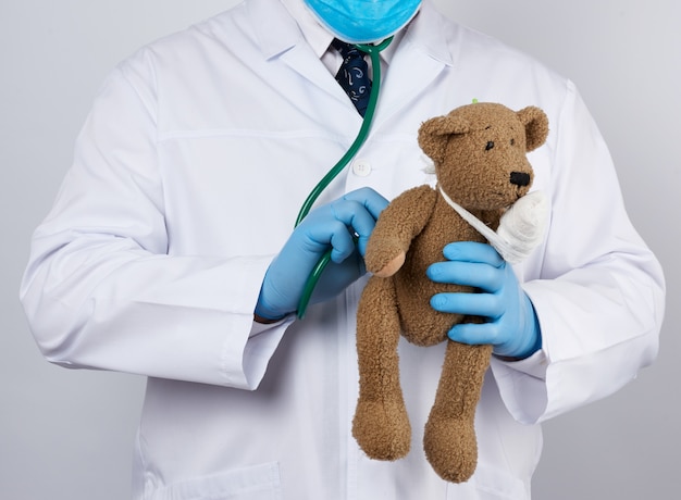 Kinderarts in een witte jas, blauwe latex handschoenen houdt een bruine teddybeer met een verbonden poot