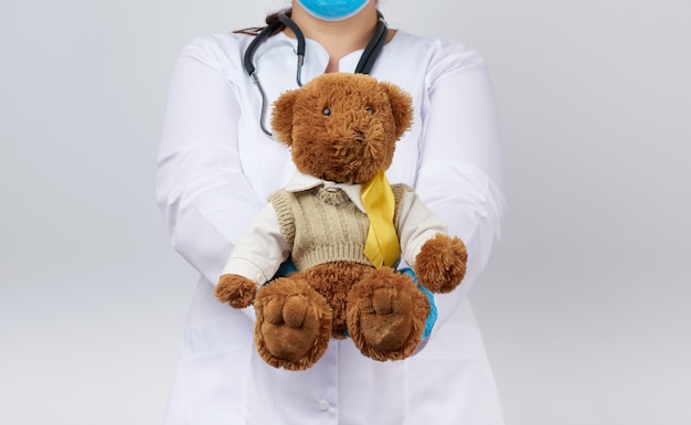 Kinderarts in witte jas, blauwe latex handschoenen houdt een bruine teddybeer met een geel lint
