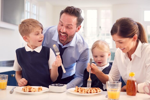 Kinderen die schooluniform dragen in de keuken en ontbijtwafels eten terwijl hun ouders zich klaarmaken voor hun werk