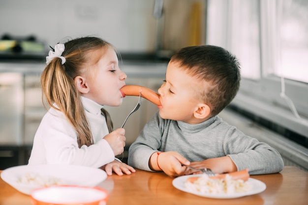 Kinderen in de keuken, jongen en meisje, eten worstjes met pasta erg leuk en delen er samen een