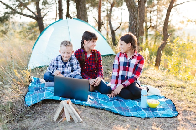 Kinderen kijken show op laptop in het bos Jongens en meisjes rusten in een tentenkamp Kinderen zitten in de zomer op de plaid op het gras