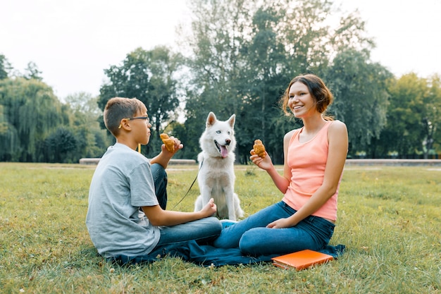 Kinderen rusten in het park op een groen grasveld met een Husky witte hond, eten croissants, praten