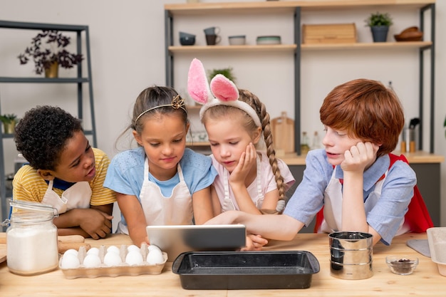 kinderen verpakt in het lezen van recepten met behulp van een digitale tablet terwijl ze aan een houten keukentafel staan om een verrassing te maken voor Moederdag