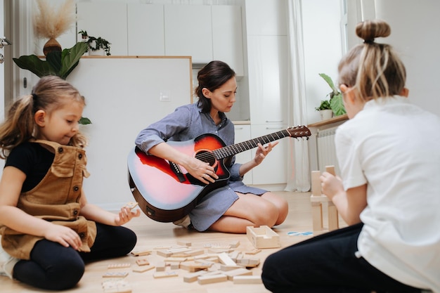 Kinderen zijn druk aan het spelen op de keukenvloer met houten blokken Terwijl moeder gitaar speelt op de achtergrond