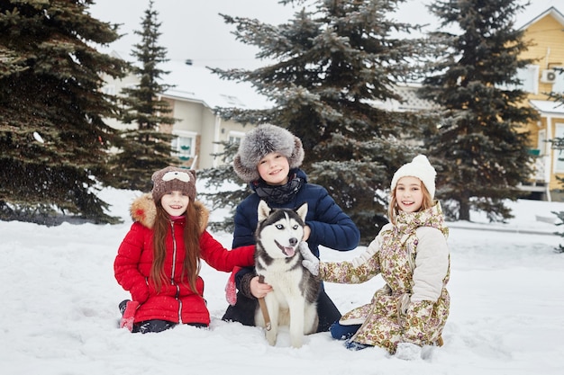 Kinderen zitten in de sneeuw en aaien hond husky. kinderen gaan in de winter naar buiten en spelen met een husky-hond. wandel in het park in de winter, vreugde en plezier, hondenhusky met blauwe ogen. rusland, sverdlovsk, 28 dec 2017