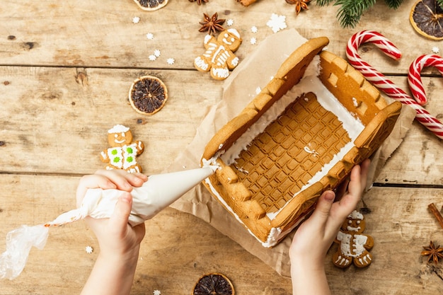 Kinderhand versieren gingerbread house met zoete glazuur. handgemaakt proces, feestelijk nieuwjaarsdecor. traditionele kruiden, houten achtergrond, bovenaanzicht