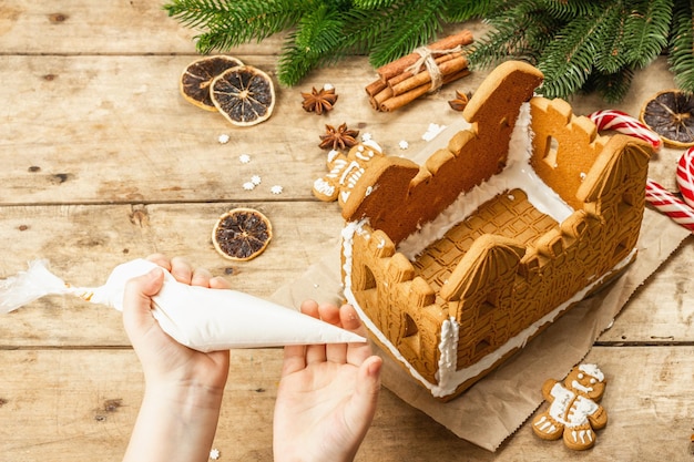 Kinderhand versieren gingerbread house met zoete glazuur. handgemaakt proces, feestelijk nieuwjaarsdecor. traditionele kruiden, houten achtergrond, close-up