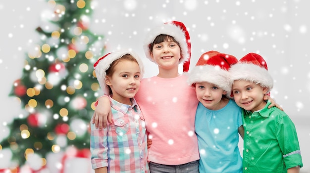 kindertijd, wintervakanties, vriendschap en mensen concept - groep gelukkige glimlachende kleine kinderen in kerstman hoeden die omhelzen over kerstboom en sneeuw