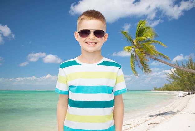 kindertijd, zomer, reizen, vakantie en mensenconcept - glimlachende kleine jongen die zonnebril over strandachtergrond draagt