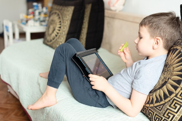 Kindjongen die op de bus zit met een tablet die spelletjes speelt of er iets op bestudeert en verse komkommer eet Sociaal en technologisch concept