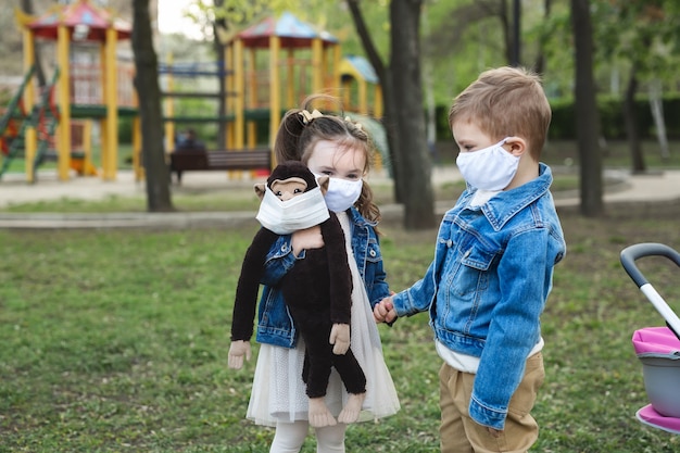 Kindjongen en meisje die in openlucht met de bescherming van het gezichtsmasker lopen. Klein meisje houdt een opgezette aap in haar handen. Coronavirus (COVID-19