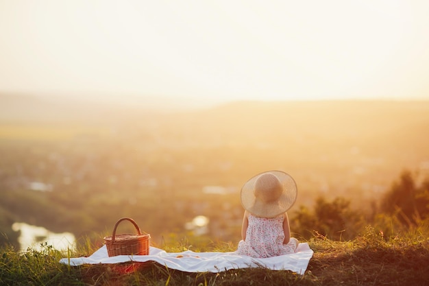 Kindmeisje in strohoed en zomerjurk zittend op de heuvel bij zonsondergang