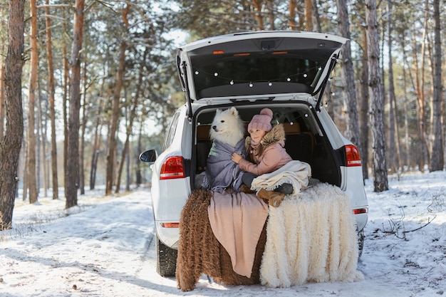 Kindmeisje zit in de kofferbak van de auto met haar huisdier, een witte hond Samojeed, in de winter in het besneeuwde dennenbos, gewikkeld in warme dekens