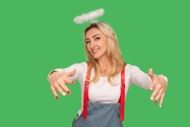 Kindness and generosity Portrait of angelic beautiful adult woman with nimbus above head holding outstretched hands and smiling giving free hugs indoor studio shot isolated on green background