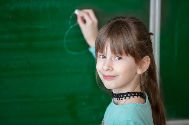 Kindstudent in de klas Het gelukkige schoolmeisje bij het groene bord schrijft iets