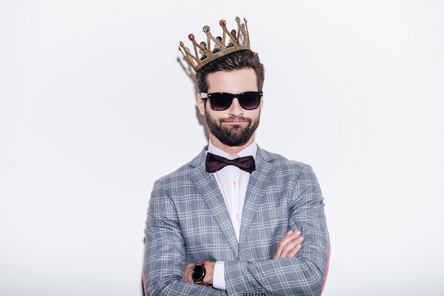 King of style. Sneering young handsome man wearing suit and crown keeping arms crossed and looking at camera while standing against white background