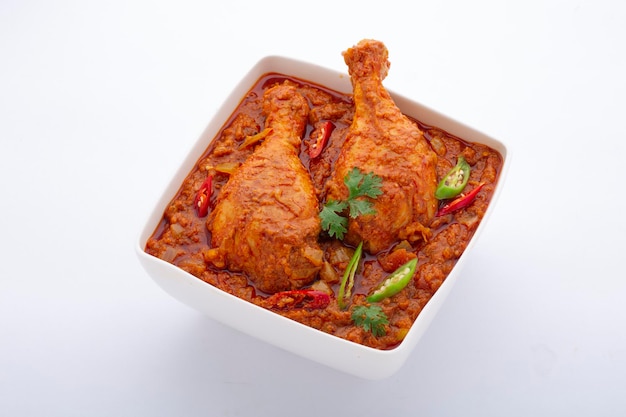 Kipcurry of masala, pittig roodachtig kippenpootstuk gegarneerd met korianderblad en verse groene chili die is gerangschikt in een witte keramische kom met witte achtergrond, geïsoleerd.