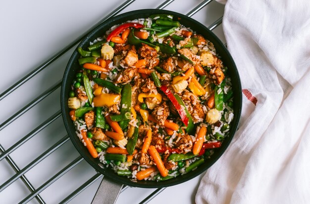 Kippenvlees, rijst en groenten in een zwarte pan op de witte tafel.