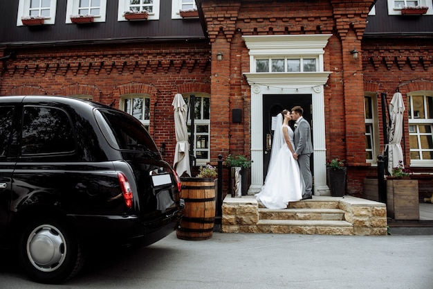 イギリスのウェディングドレスを着た車の横にある新婚の新郎新婦のキス。美しい新婚夫婦