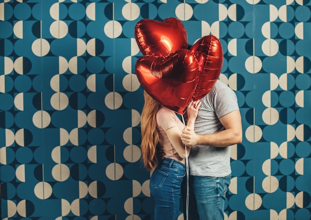 Kissing caucasian couple are hiding behind some air heart sharped balloons on a blue  wall