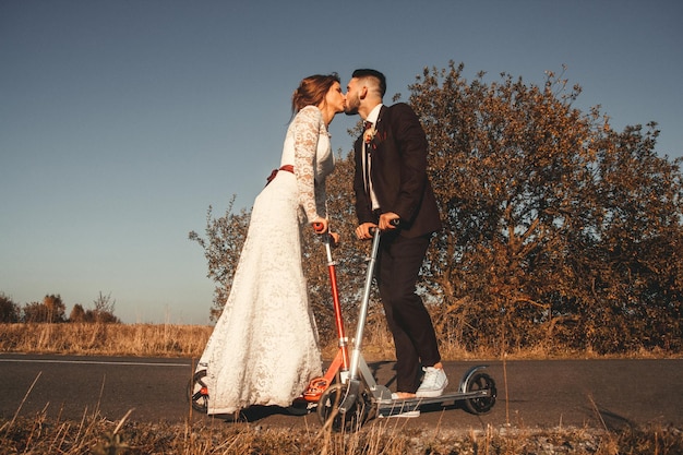 Kissing wedding couple riding a on scooters along the road outside the city at sunset