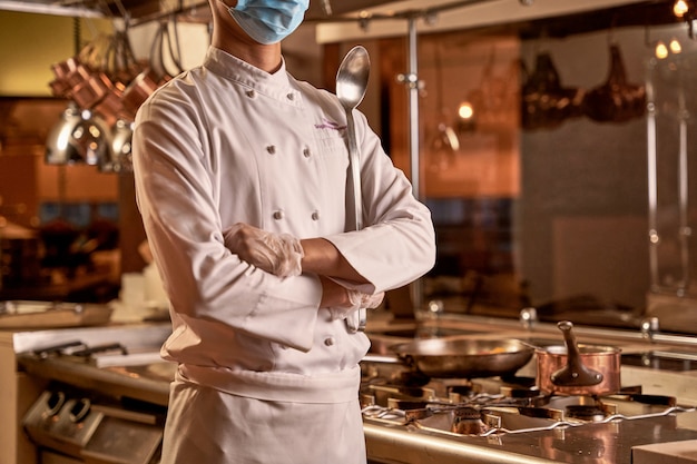 Kitchen employee crossing hands below chest while staying beside a cooker with pot and frying pan