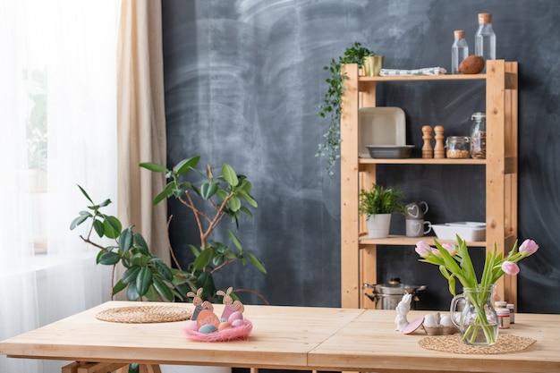 Kitchen interior with Easter decorations and flowers in vase on table, crockery on shelves against blackboard