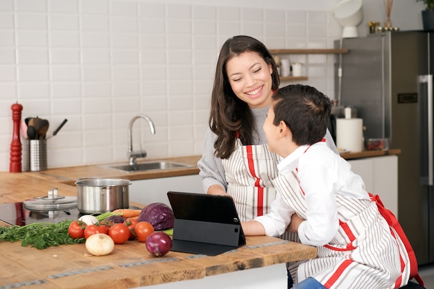 In the kitchen a son searches for a cooking recipe online via his tablet children helping parents he...