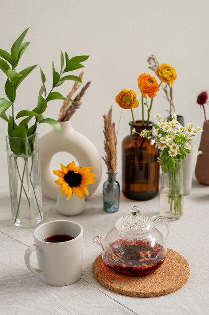 Tavolo da cucina con tazza bianca in porcellana, teiera con tè nero, gruppo di fiori vari e fiori di campo essiccati in vasi in piedi sulla parete