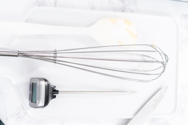 Kitchen whisk and candy thermometer on a white cutting board.