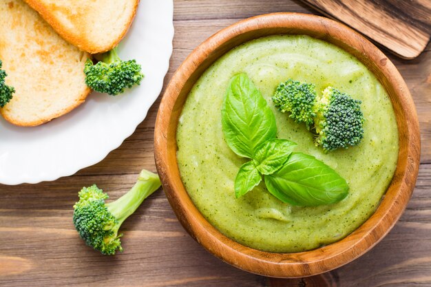 Klaar om verse hete broccolipureesoep met stukken broccoli en basilicumbladeren te eten in een houten plaat op een houten lijst.