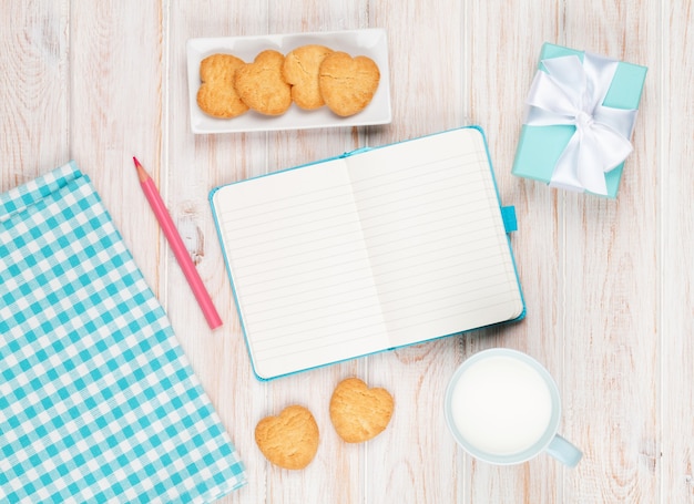 Kladblok, kopje melk, hartvormige koekjes en geschenkdoos op witte houten tafel