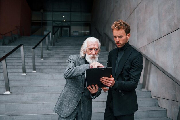 Kladblok vasthouden Jonge kerel met senior man in elegante kleding is buiten samen Conceptie van zaken