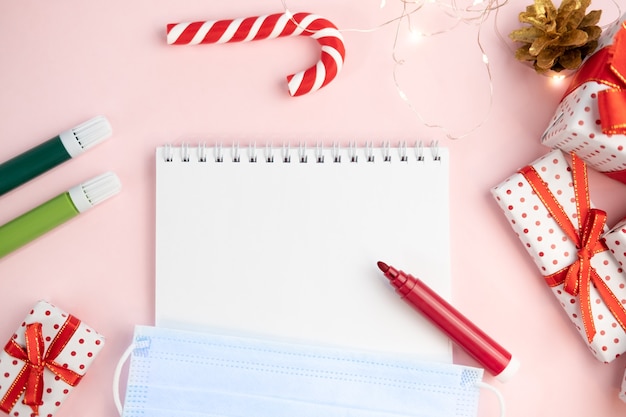 Kladblok-verlanglijstje op een roze tafel met viltstiften met beschermend gezichtsmasker op een kerstachtergrond. Concept van kerstmis, nieuwjaar, plannen en wensen maken en coronavirus