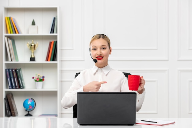 Klantenservice schattig blond meisje office shirt met headset en computer wijzend op cup