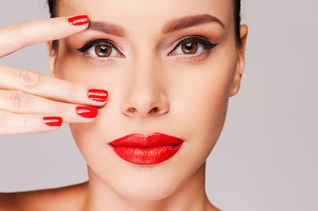 Klassiek rood. Close-up van een mooie vrouw met rode manicure die vingers op het gezicht houdt terwijl ze tegen een grijze achtergrond staat