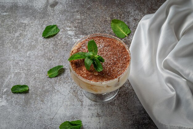 Foto klassiek tiramisu-dessert met muntblaadjes in een glas, met cacao en chocolade op de achtergrond van een grijze stenen tafel