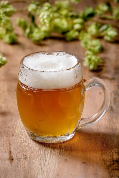 Klassieke glazen mok vers koud schuimend lagerbier met groene hopbellen erachter over houten tafel. Kopieer ruimte