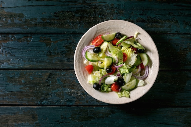 Klassieke groentesalade met tomaten, komkommer, ui, slablaadjes en zwarte olijven in witte keramische plaat. donkere houten achtergrond. plat leggen, kopie ruimte