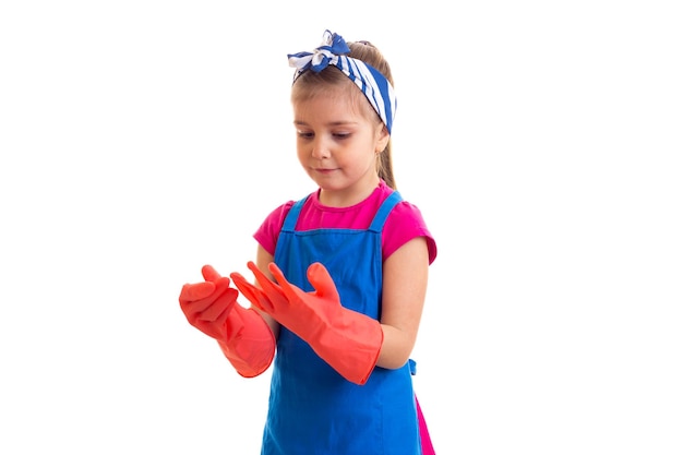 Klein aardig meisje met paardenstaart in roze shirt en blauwe schort met rode handschoenen in studio