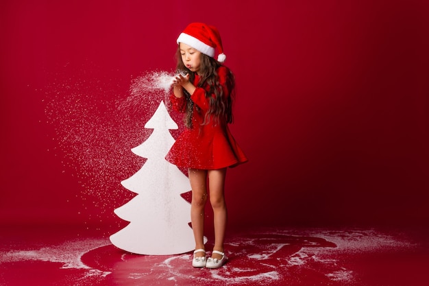 klein Aziatisch meisje in een kerstmuts blaast de sneeuw uit haar handpalmen kerstconcept tekstruimte
