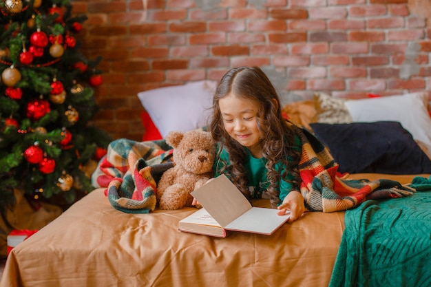 Klein aziatisch meisje in pyjama in de slaapkamer liggend op bed bij de kerstboom die een boek leest