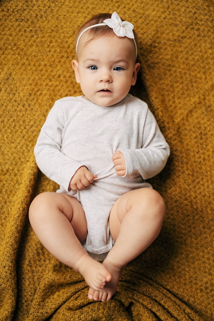 Klein babymeisje met een bloem op haar hoofd in een witte bodysuit ligt met haar handen op een gele deken