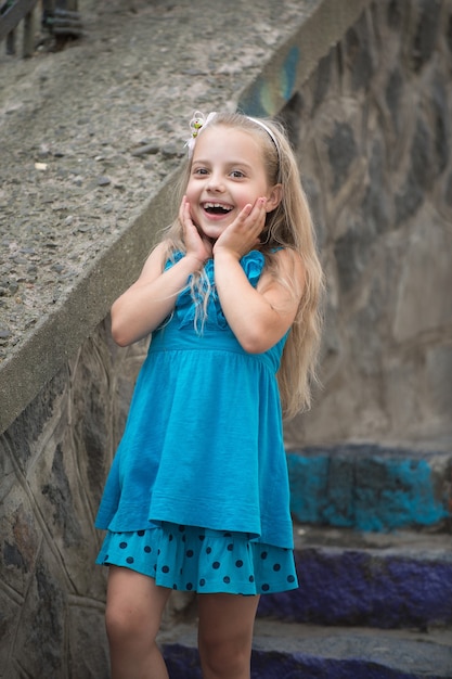 Klein babymeisje of schattig gelukkig kind met schattig lachend verrast gezicht en boog in blonde haren in blauwe jurk in de zomer buiten in de buurt van steenachtige trappen op onscherpe achtergrond