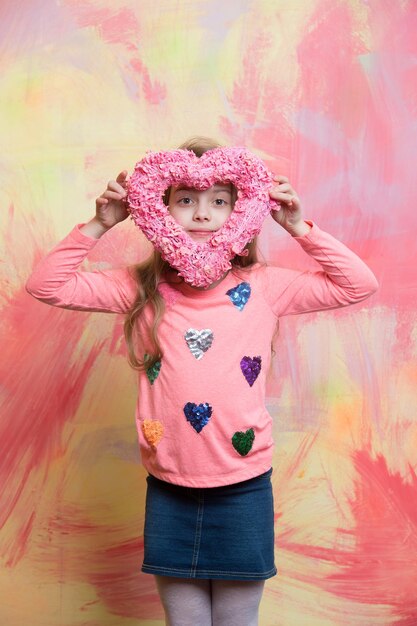 Foto klein babymeisje of schattig kind met blij gezicht en blond haar in shirt en rok met roze valentijnsdag vakantie decoratief hart op kleurrijke abstracte achtergrond