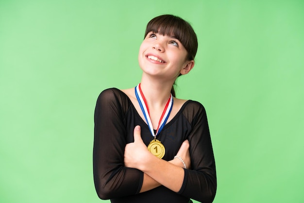 Klein blank meisje met medailles over geïsoleerde achtergrond die omhoog kijkt terwijl ze glimlacht