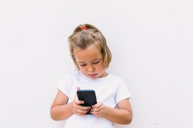 Klein blond haarmeisje met wit t-shirt, kijkend naar mobiele telefoon, met geconcentreerd gezicht