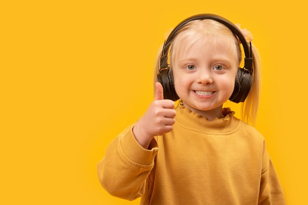 Klein blond meisje draagt een zwarte koptelefoon glimlachend en toont thumbs up Portret van preschool meisje op gele achtergrond Kopieer ruimte