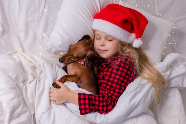 klein blond meisje in een kerstmuts en pyjama slaapt in bed met haar geliefde hond teckel