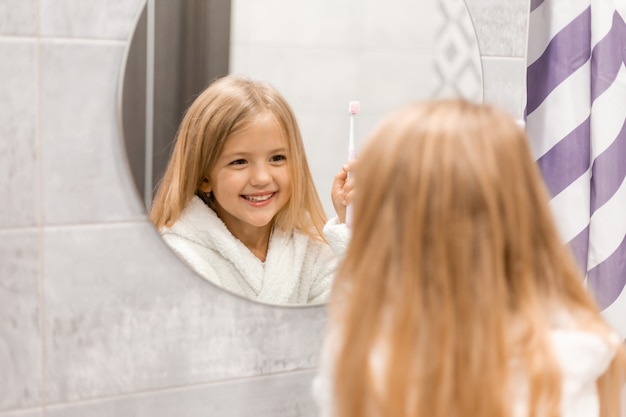 klein blond meisje in een witte badjas poetst haar tanden voor de badkamerspiegel